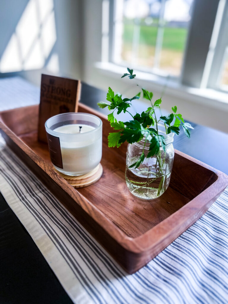 clean and clutter free black rectangle dinner table with a grey pin-striped and white table runner with a wooden centerpiece holding a candle, a mason jar with green herbs in it, and a wooden small piece of art and a beautiful window in the background