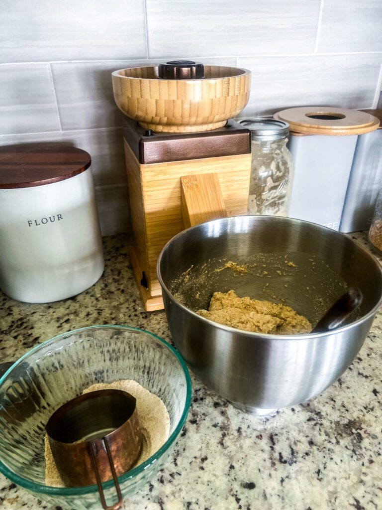 nutrimill harvest grain mill in bronze with a stainless steel bowl of dough in front of it and a canister of flour next to it, a sourdough starter in a glass mason jar, and a jar of wheat berries