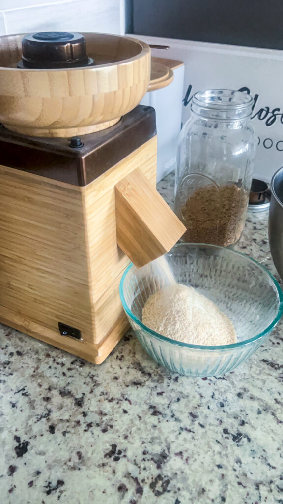 nutrimilli grain mill in bronze milling fresh whole wheat flour into a bowl with wheat berries in a mason jar in the background