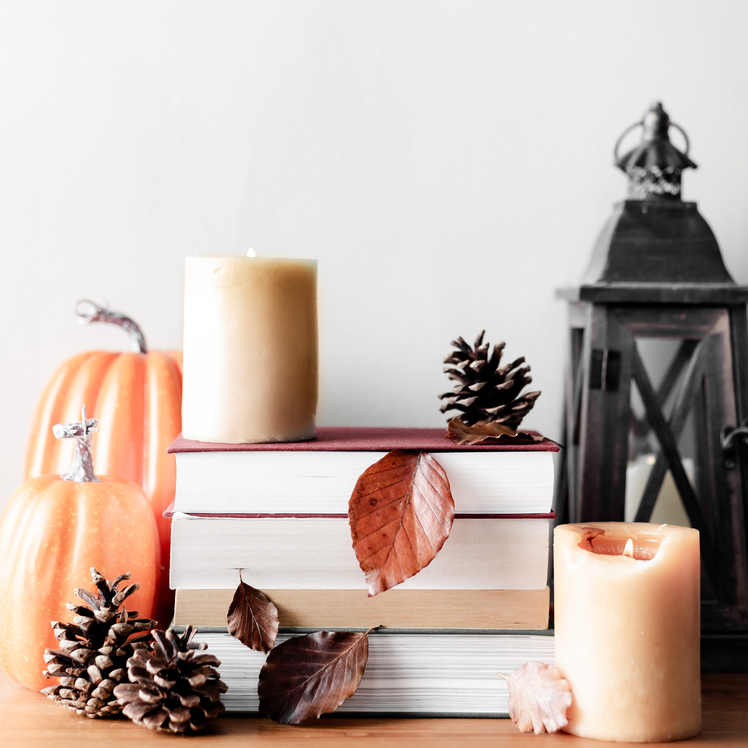 candles lit by a stack of books with fall decor around it like a black lantern, pine cones, leaves, and orange pumpkins