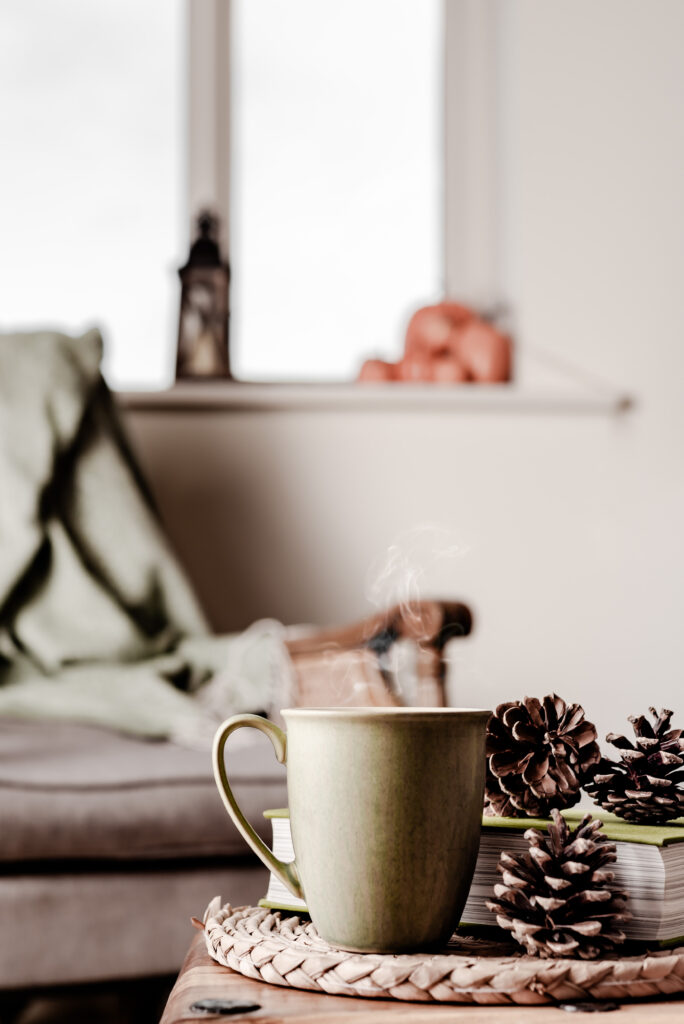  photo of a cozy couch with an olive green blanket draped over it and in focus is an olive green coffee mug with steaming coffee on a wooden table next to fall decor like pine cones
