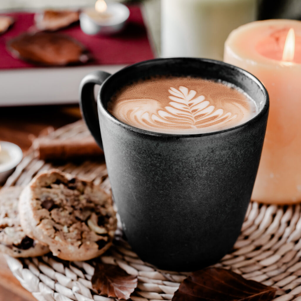 a black mug with coffee in it with a coffee art leaf on a straw placemat with a lit candle near it and a chocolate chip cookie