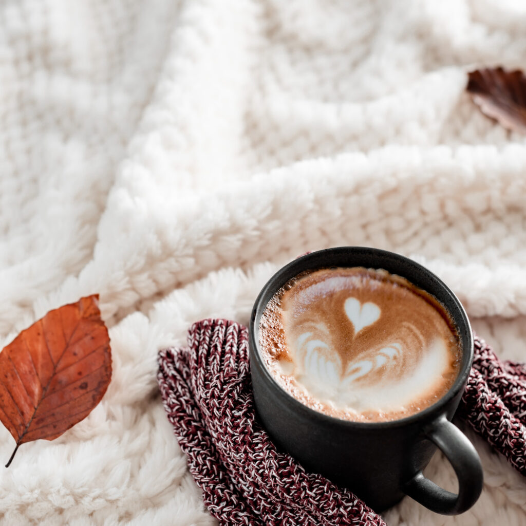 black coffee mug full of coffee with coffee ar hear in it on top of a spice colored cloth that is on top of a cozy white blanket and a decorative brown leaf