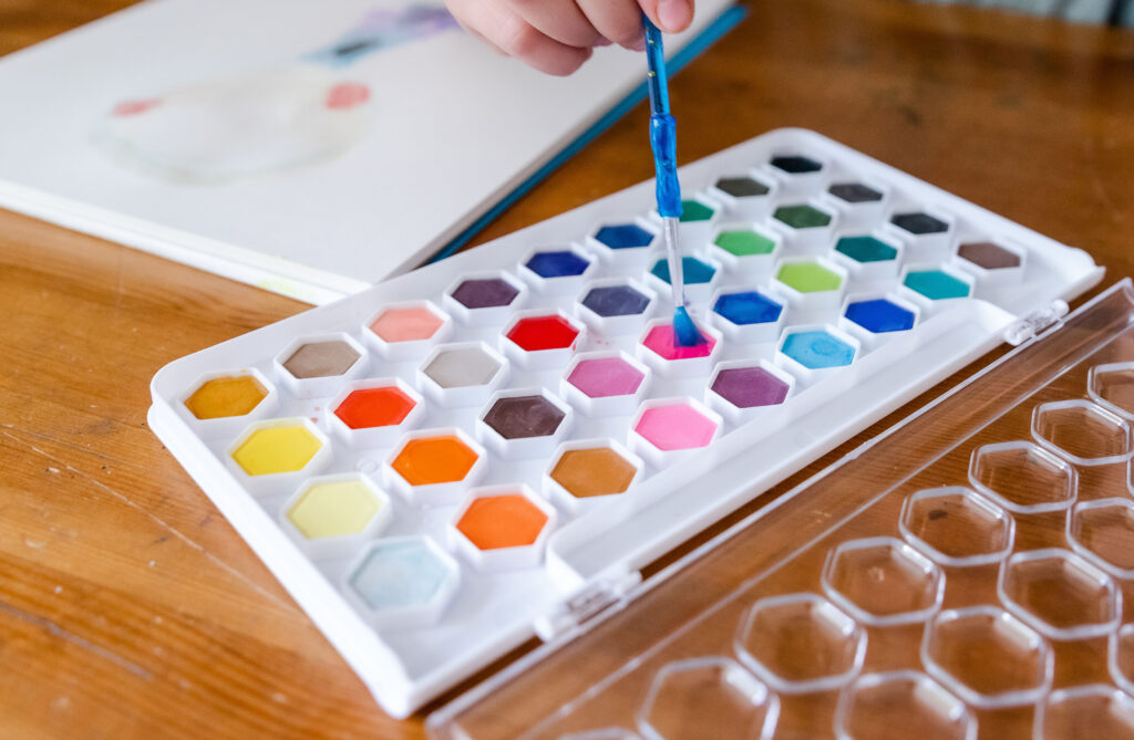 a watercolor paint palette on a wooden table with a hand dipping a paint brush into a pink color and a paper in the back ground