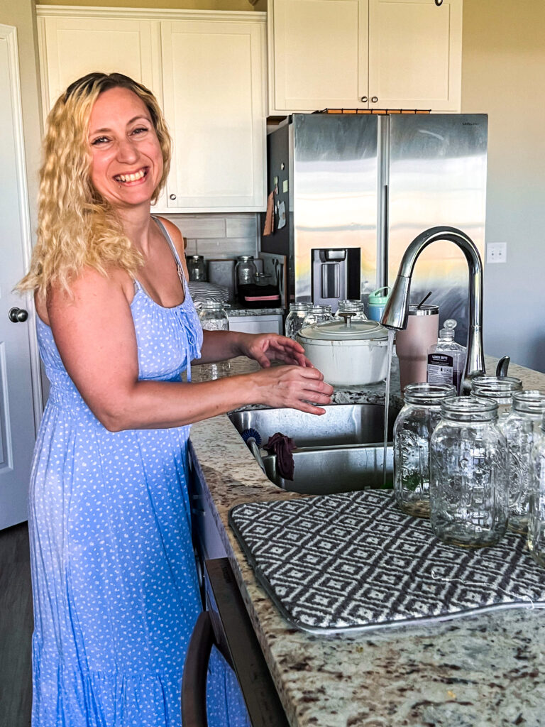 a blonde lady in a blue dress at the sink in her island washing and preparing glass mason jars