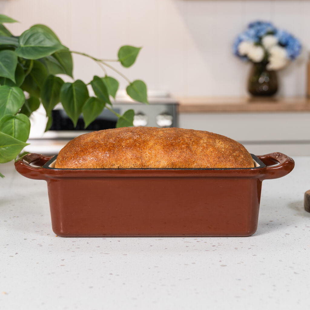 nutrimill enameled cast iron bread pan in cinnamon color with a homemade loaf of bread in it sitting on top of a countertop with a plant near it and a bouquet of flowers in the background