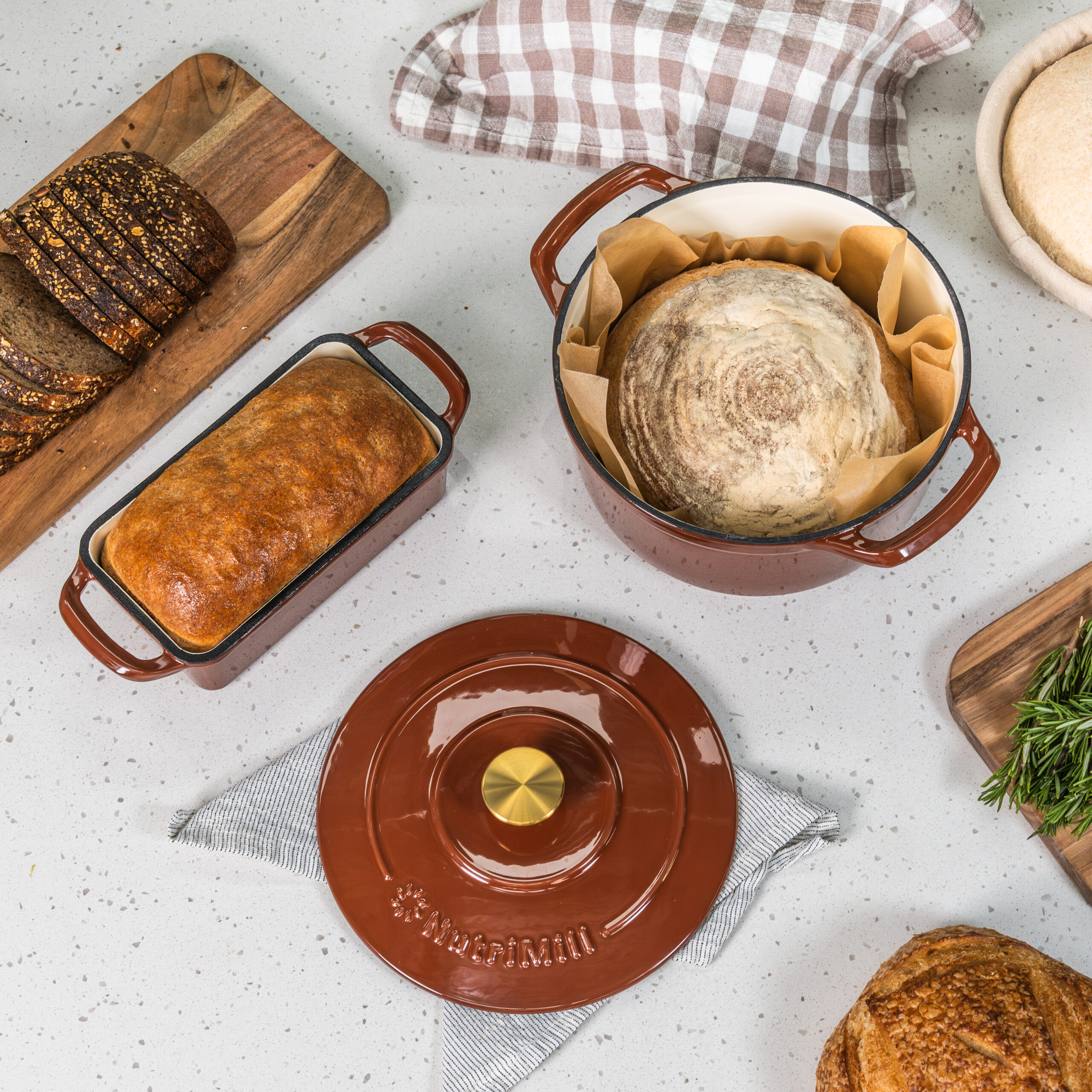 enameled cast iron dutch oven with sourdough bread in it with parchment paper the lid with a brass knob on the white countertop in front of it and the matching enameled cast iron bread pan with a homemade loaf next to it