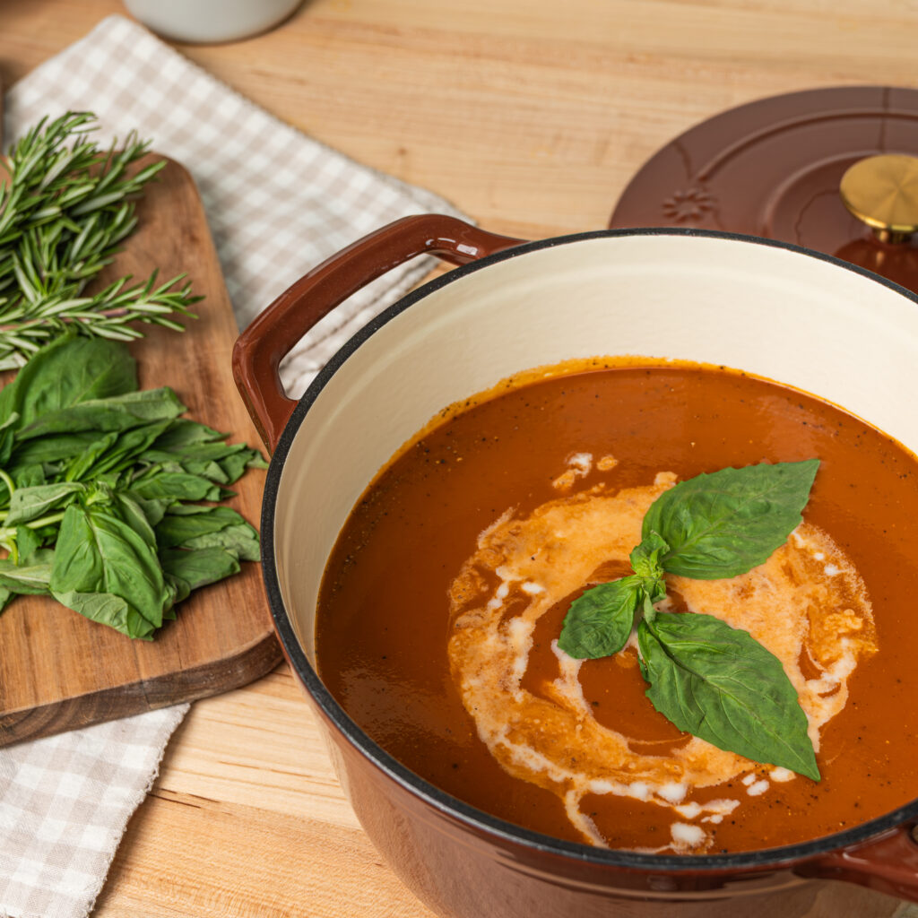 tomato colored soup topped with fresh basil in a dutch oven on top of a wooden countertop with a wooden cutting board in the background that has fresh basil on it and a checkered cloth underneath