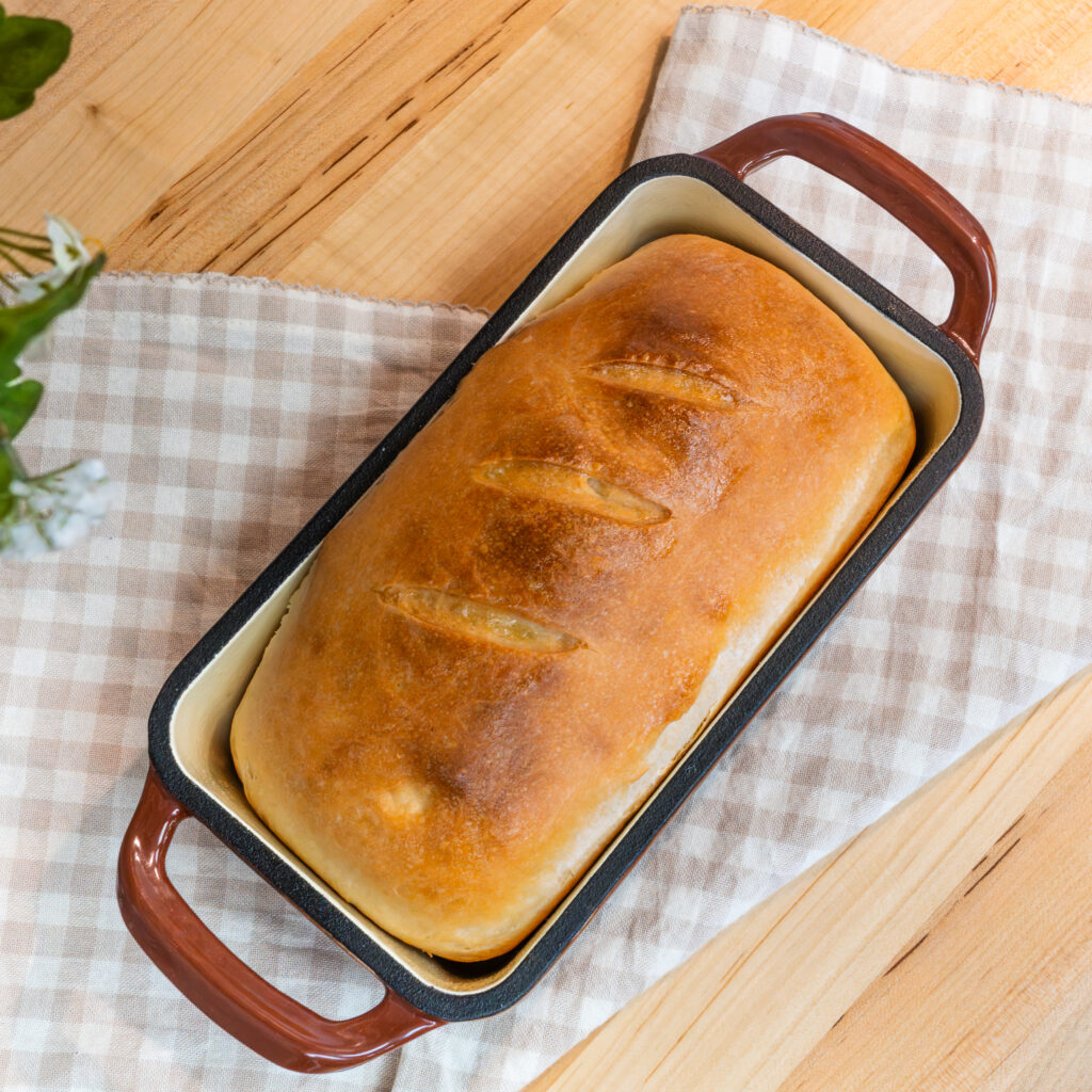 a loaf of homemade bread in an enameled cast iron bread pan on top of a checkered cloth on top of a wooden countertop