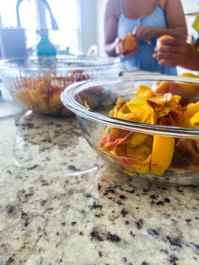 a glass bowl of peach pieces next to another glass bowl of peach peels and skins on top of a granite counter top with hands of peaches behind