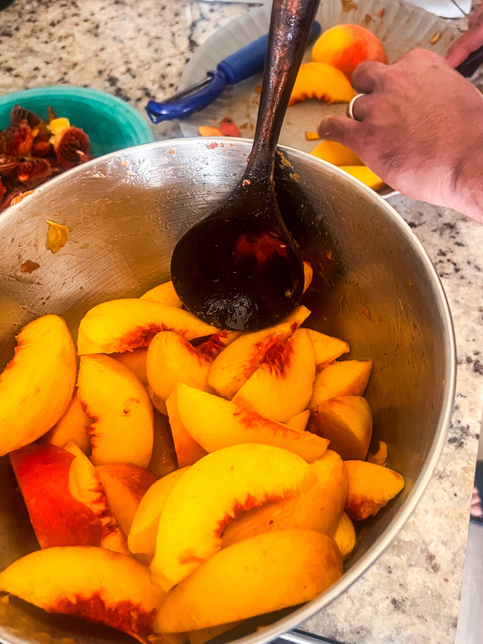 a stainless steel bowl full of peach slices and lemon juice with a wooden spoon in it and a man's hands in the background slicing peaches