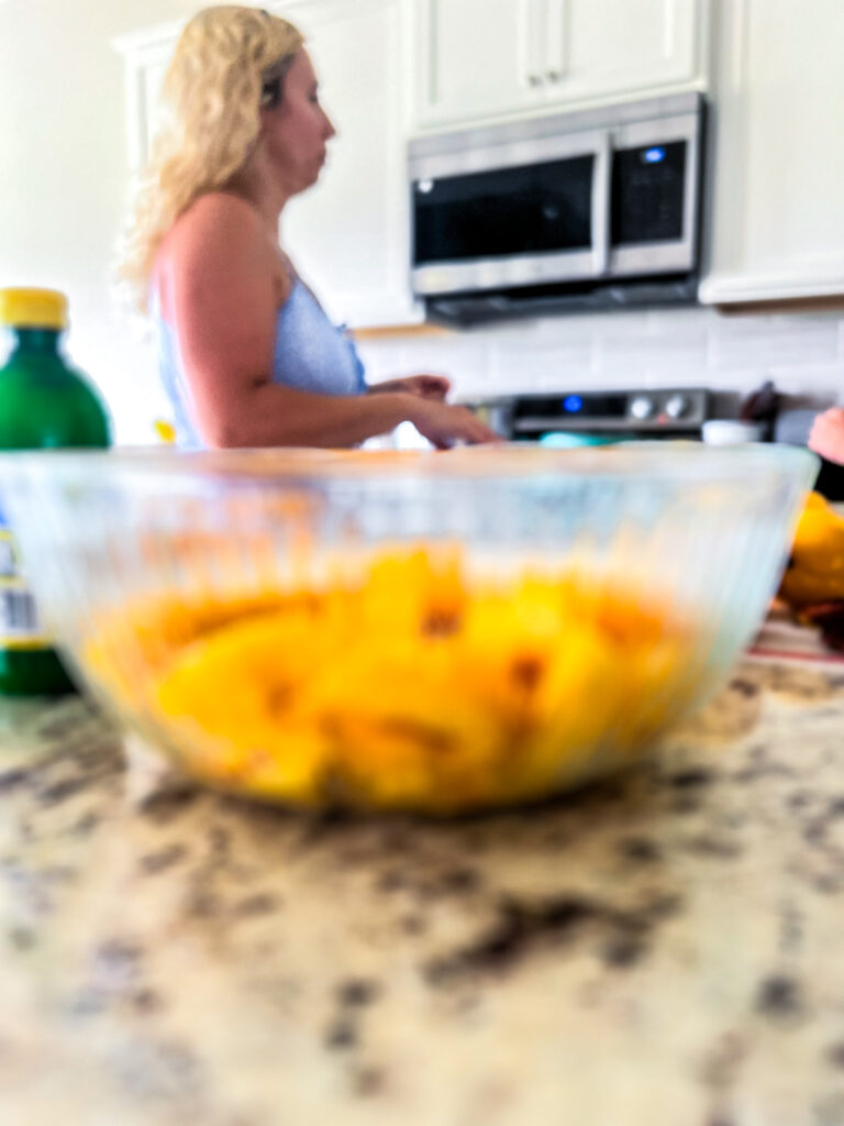 a glass bowl of sliced peaches on a granite countertop with a blonde lady in a blue dress standing at her stove