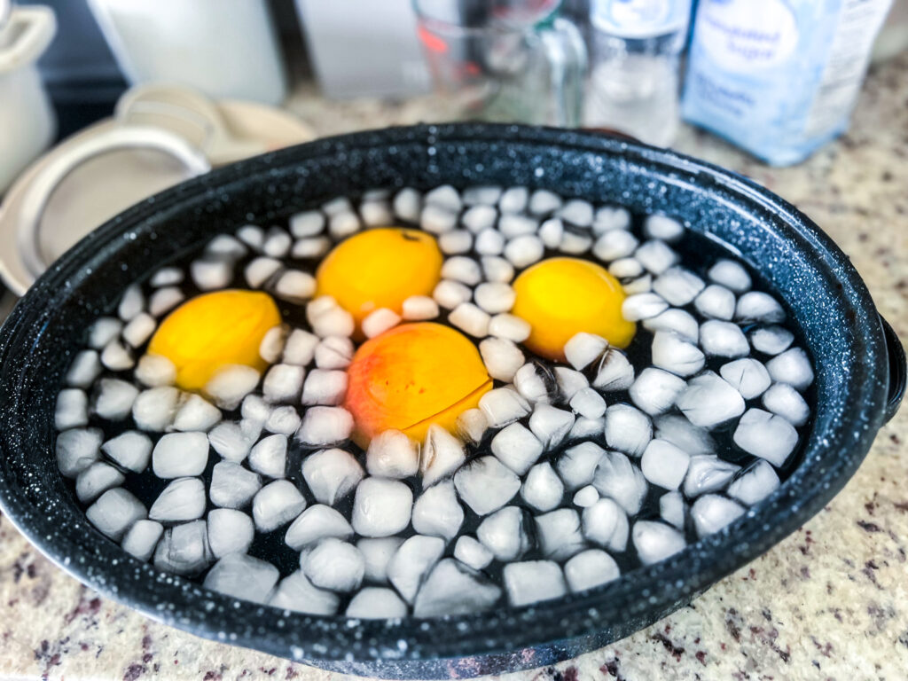 four blanched peaches sitting in an ice bath in a large black roasting pan on top of a counter
