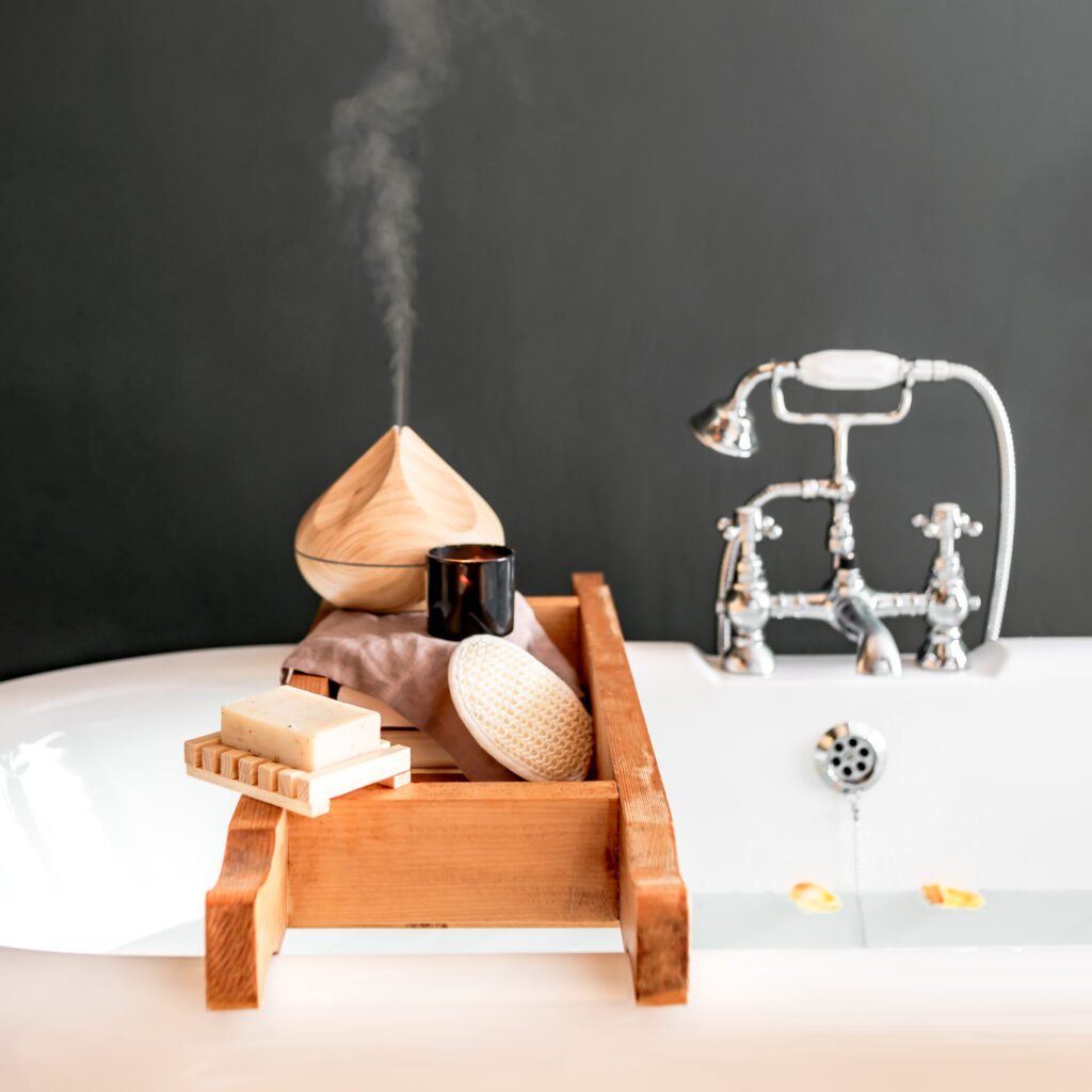 A vintage white bathtub with a vintage sliver spout and handle that has a wooden spa tray on it holding soap and a diffuser and other self care items