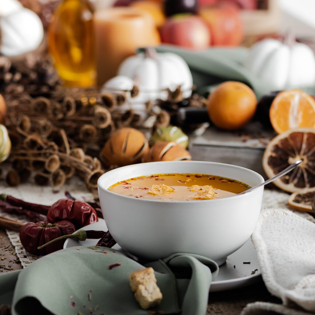 a white bowl of steaming hot soup on a black table with fall decor all around it  like small orange pumpkins, white pumpkins, pine cones, and dried oranges