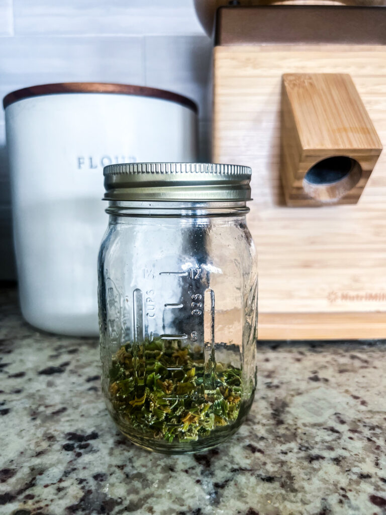 different angle 1/2 cup of dehydrated celery in a close mason jar on top of a granite countertop with a flour canister and a grain mill in the background