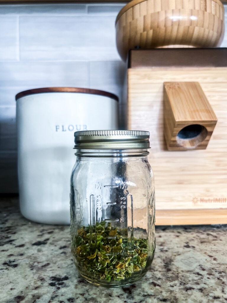 1/2 cup of dehydrated celery in a close mason jar on top of a granite countertop with a flour canister and a grain mill in the background