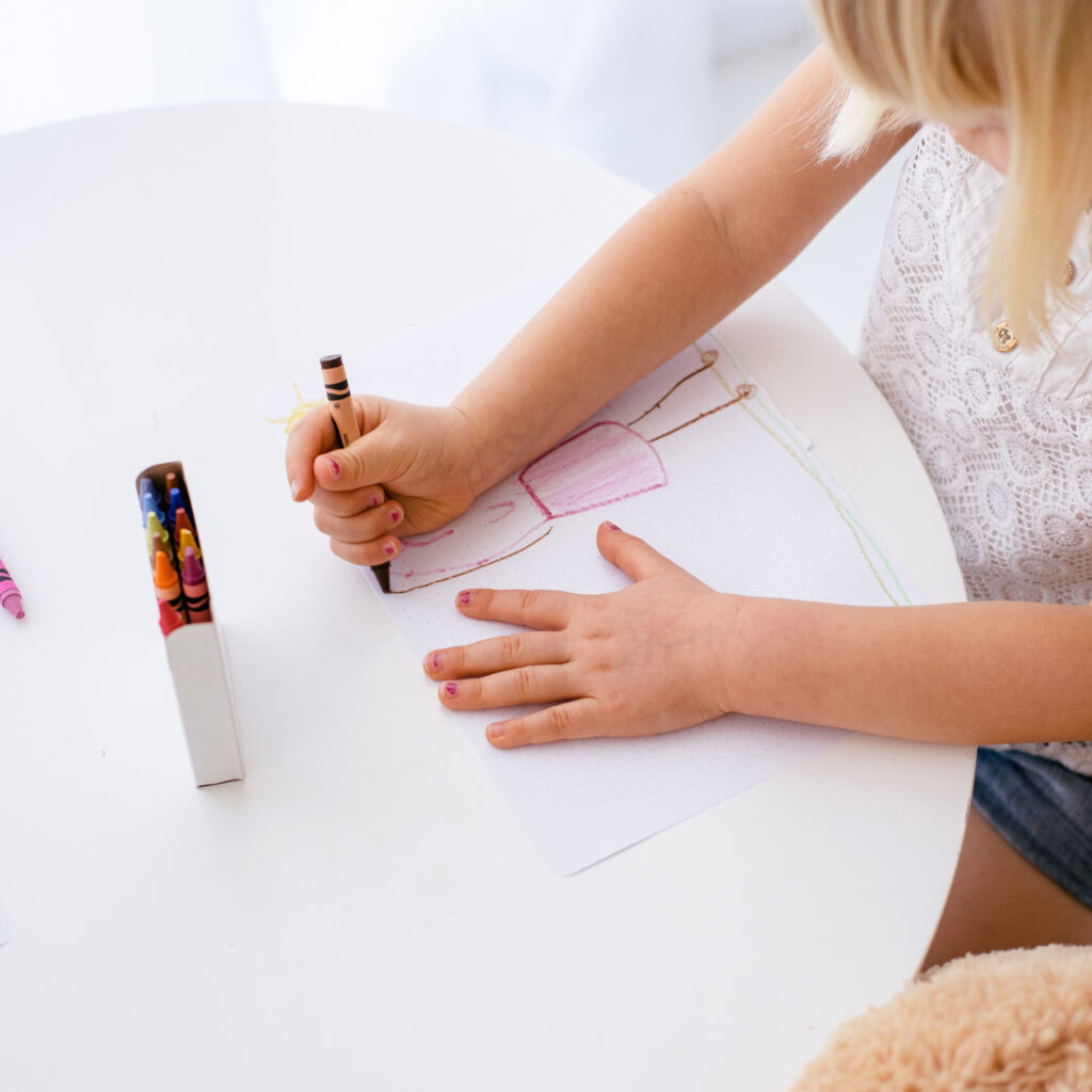 a child's hands drawing a person on a white piece of paper on top of a white table with a box of crayons