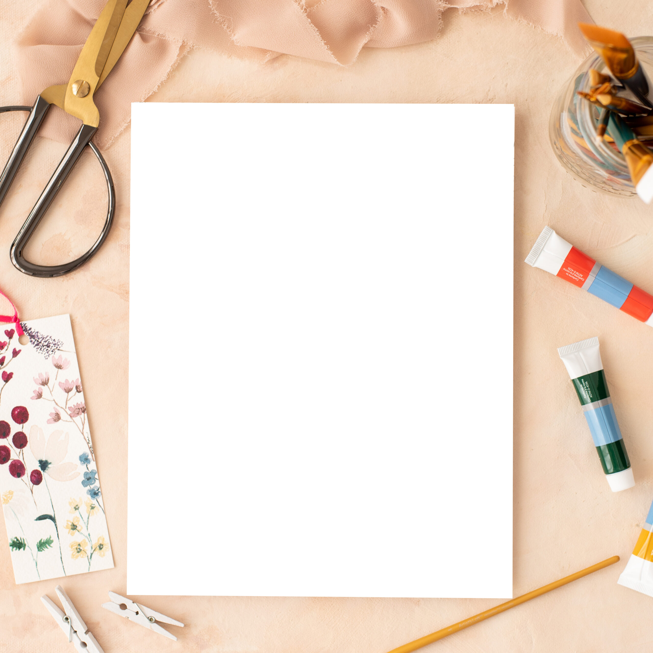 a blank white paper on a peach table with paint tubes and paint brushes next to it and scissors and patterned fabric