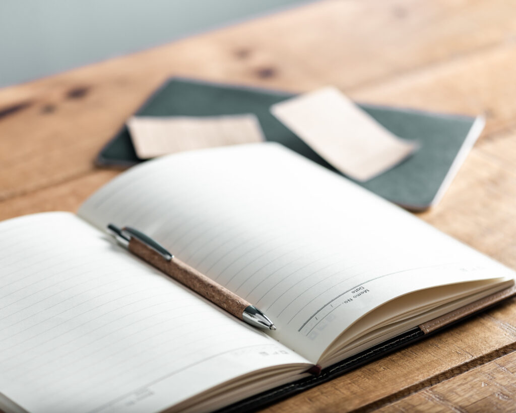 a pen on an open notebook on top of a wooden table with a close notebook and sticky notes in the background
