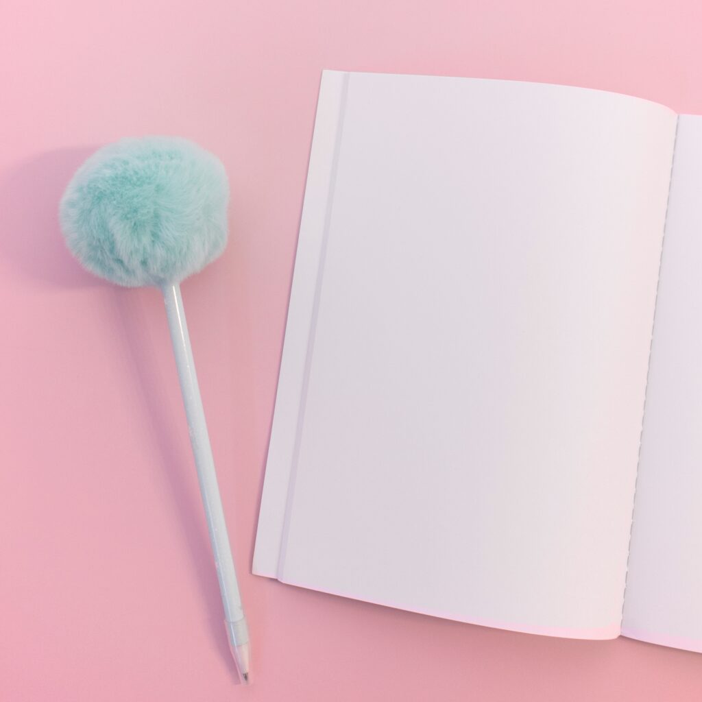 a blank white book and white pen with fuzzy blue end on top of a light pink table