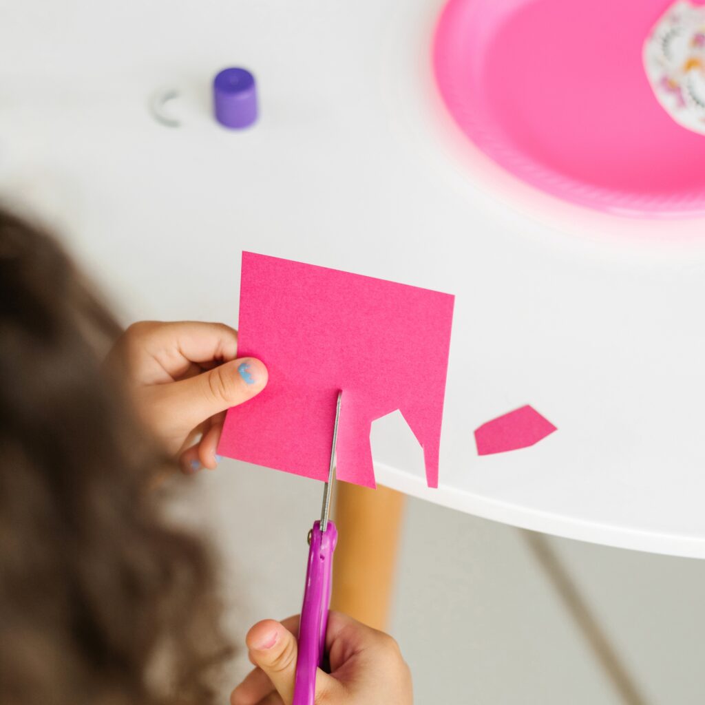 a child cutting pink paper with pink scissors and in the background is a white table with a pink plate and more craft supplies