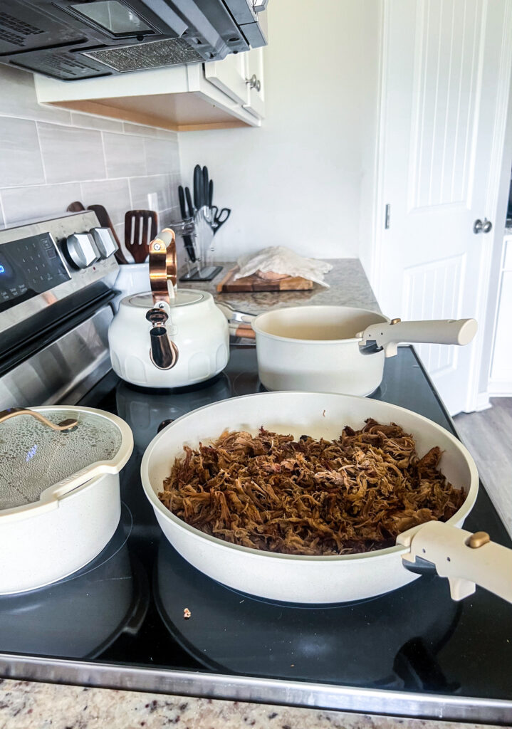 skillet of shredded pork on top of a stove with other cream colored cookware on the stove