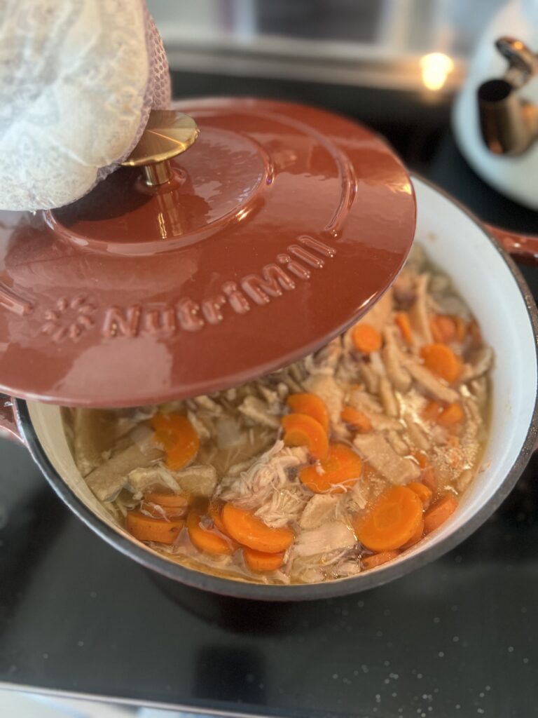 cinnamon colored Nutrimill non-toxic enameled cast iron dutch oven with homemade chicken noodle soup inside; homemade freshly milled flour noodles, carrots, chicken, and more, with a hand with a mitt holding the lid to the side