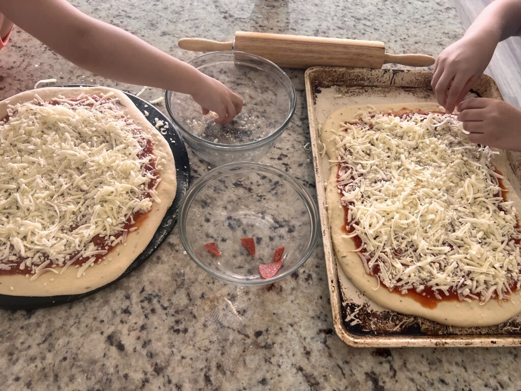 two hands of kids topping two homemade pizzas with mozzarella cheese on top of a granite countertop with a wooden rolling pin in the background