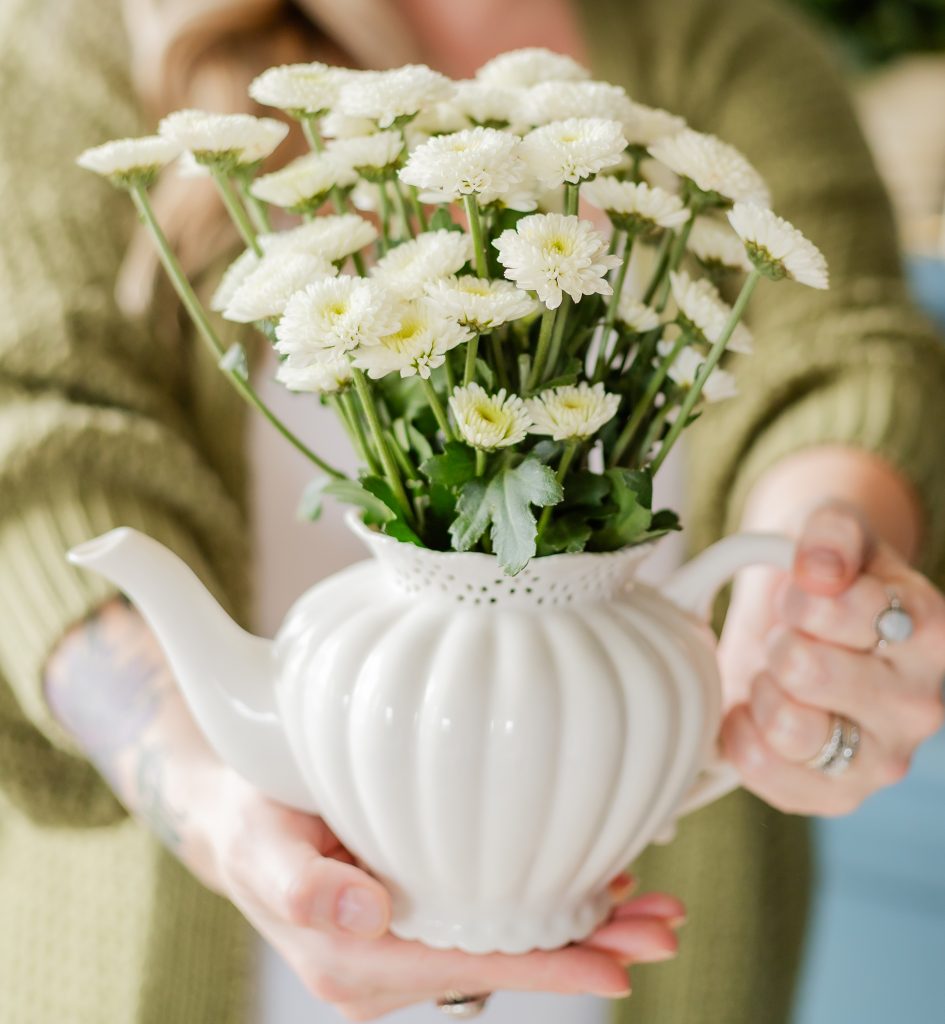 A white porcelain teapot vase filled with small white flowers held by a lady in a green cardigan