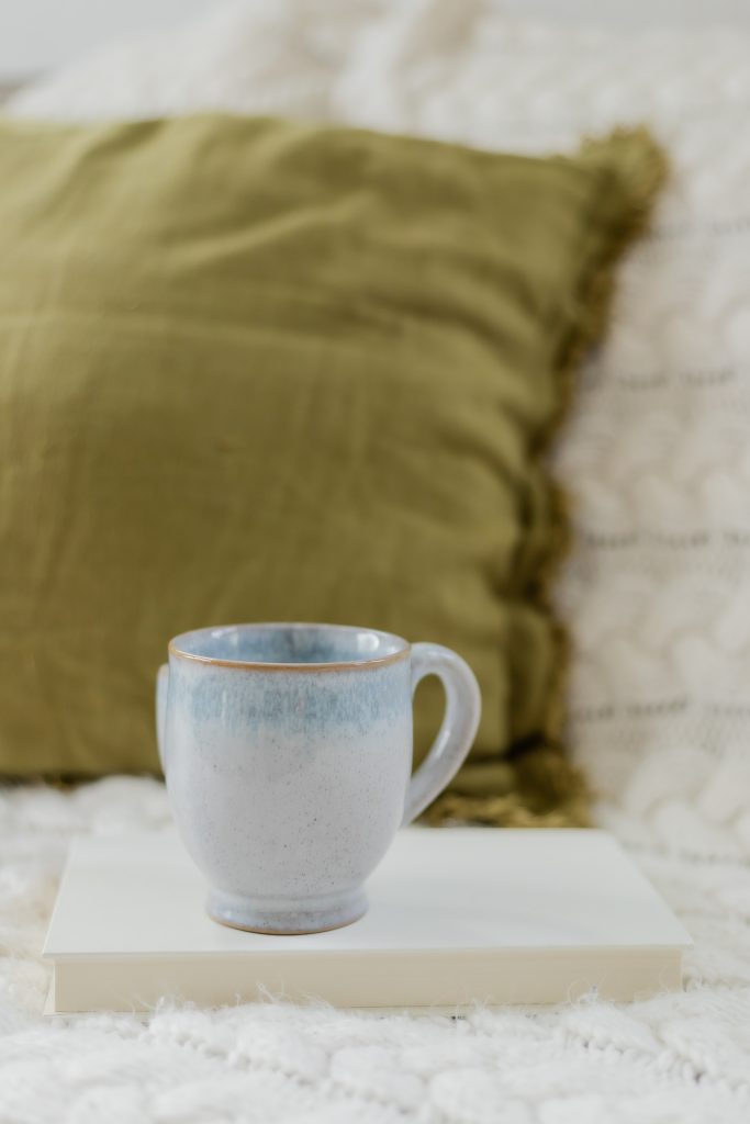 blue coffee mug on top of a book on top of a cozy white quilt with an army green pillow